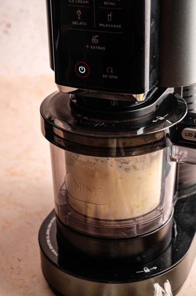 Process shot of a Ninja Creami machine blending a creamy pineapple and yogurt mixture in a clear container.