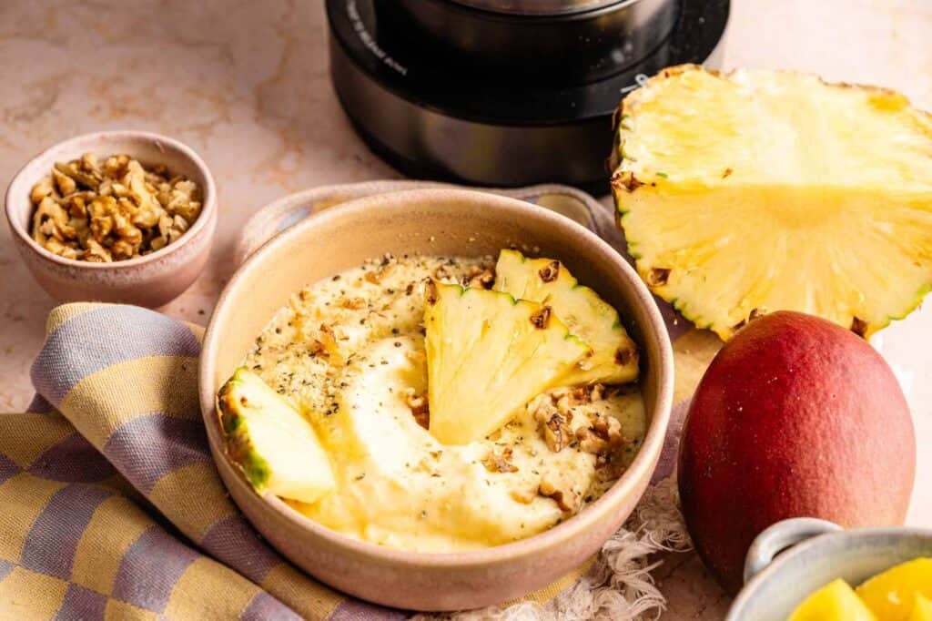 Ninja Creami smoothie bowl topped with fresh pineapple slices, chopped nuts, and seeds, served in a ceramic bowl with mango and pineapple on the side.