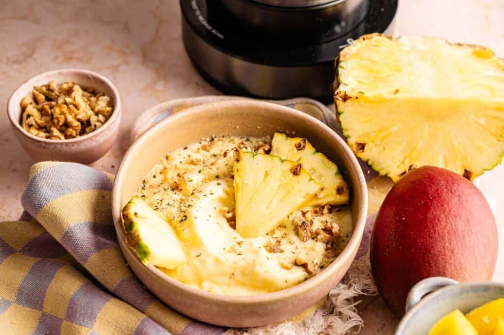 A Ninja Creami smoothie bowl topped with three pineapple slices and crushed walnuts, served with a side bowl of extra walnuts, a whole mango, and a half pineapple in the background.