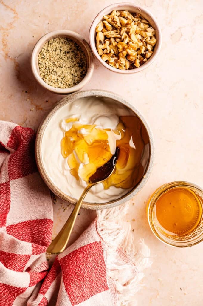 Ninja Creami frozen yogurt ingredients: Greek yogurt with honey being stirred by a gold spoon, hemp seeds, and chopped walnuts on a marble surface.