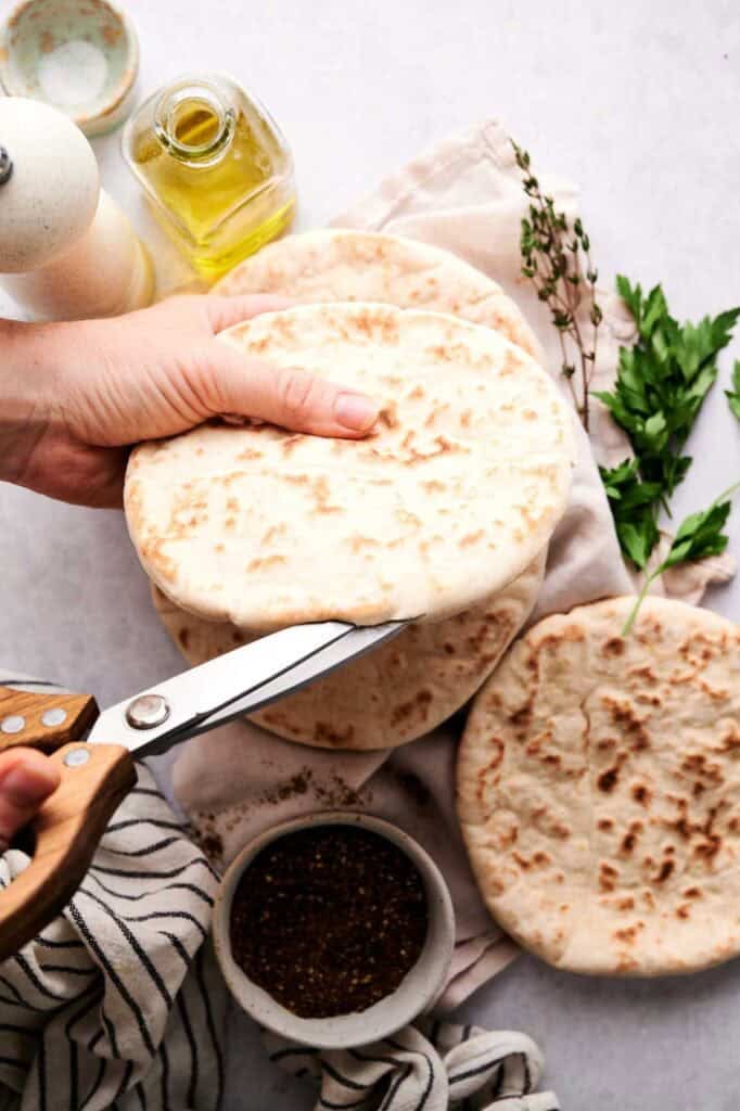 Hands holding stacked pita breads with scissors ready to cut, surrounded by olive oil, za'atar, and fresh herbs.