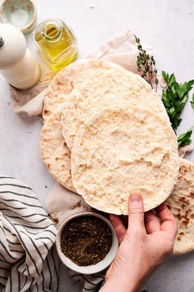 Hand separating a pita bread in half, with a bowl of za'atar, olive oil, and fresh herbs nearby.