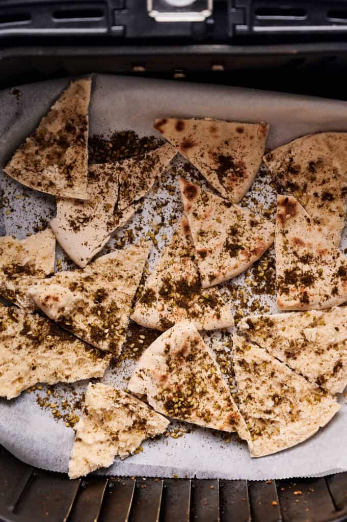 Triangular Air Fryer pita chips seasoned with herbs and spices, arranged on parchment paper inside an air fryer basket with scattered seasoning crumbs.