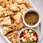 A white oval platter with air fryer pita chips seasoned with za’atar, served alongside a small bowl of za’atar spice mix and a fresh chopped cucumber-tomato-parsley salad.