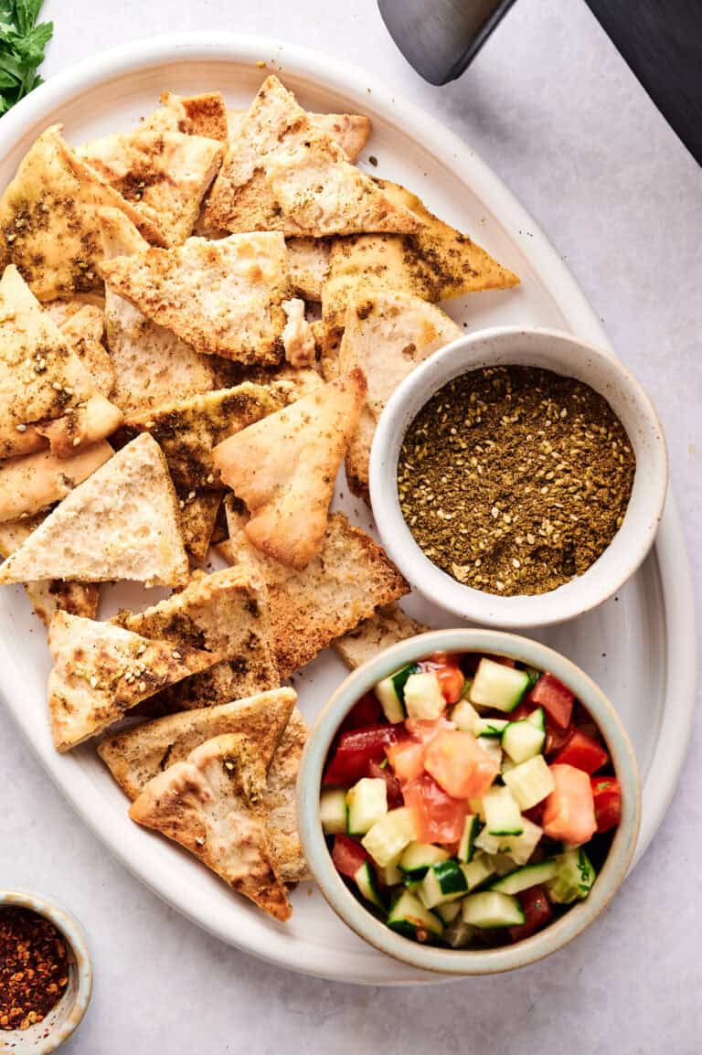 A white oval platter with air fryer pita chips seasoned with za’atar, served alongside a small bowl of za’atar spice mix and a fresh chopped cucumber-tomato-parsley salad.