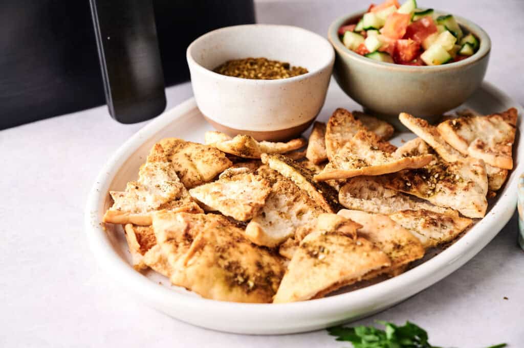 Za'atar-seasoned air fryer pita chips on a white oval platter, served alongside a bowl of za'atar spice and a fresh cucumber tomato salad.