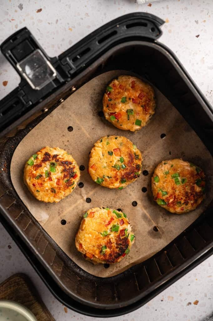 Five golden-brown air fryer salmon croquettes with diced red and green bell peppers, arranged on perforated parchment paper inside a black air fryer basket.