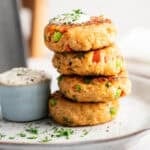 A stack of four golden-brown air fryer salmon croquettes speckled with diced red peppers and green onions, topped with a dollop of creamy sauce and fresh parsley, served on a white plate with a side ramekin of dipping sauce.