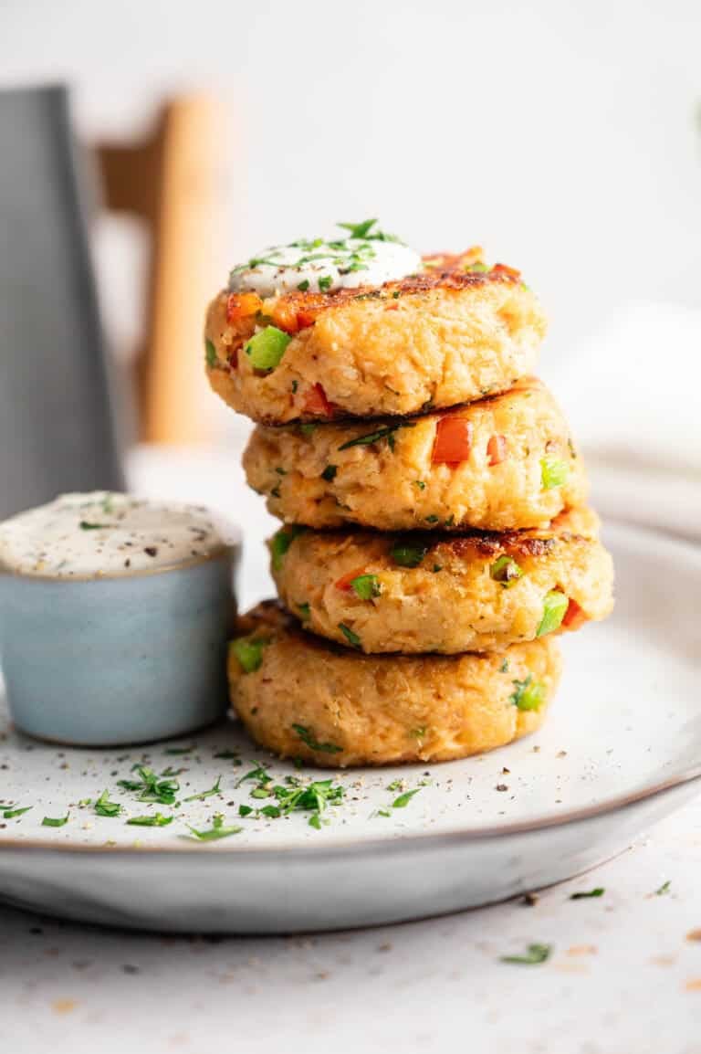 A stack of four golden-brown air fryer salmon croquettes speckled with diced red peppers and green onions, topped with a dollop of creamy sauce and fresh parsley, served on a white plate with a side ramekin of dipping sauce.