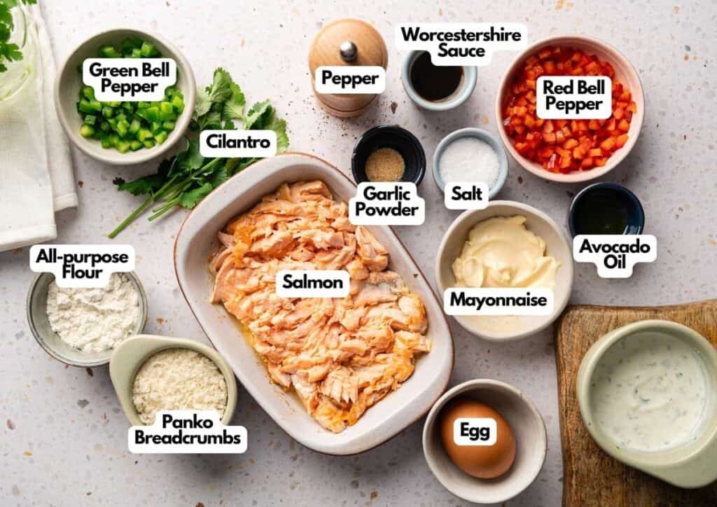 A tray of flaked salmon for air fryer salmon croquettes surrounded by prepped ingredients including diced green and red bell peppers, cilantro, flour, panko breadcrumbs, egg, mayonnaise, Worcestershire sauce, avocado oil, garlic powder, salt, and pepper on a light countertop.