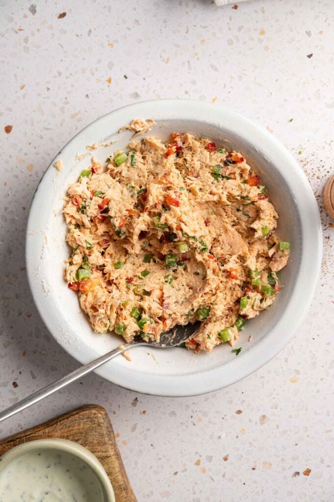Salmon croquette mixture with flaked salmon, diced red and green bell peppers, and fresh herbs in a white ceramic dish with a spoon.