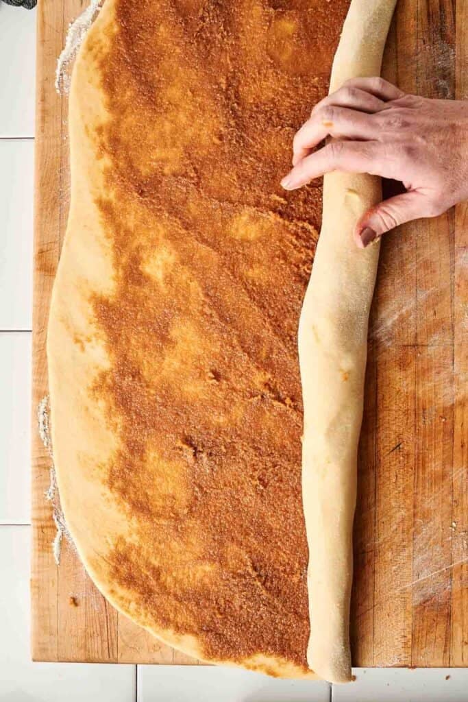 Hands rolling sourdough cinnamon roll dough spread with cinnamon sugar filling into a log on a floured wooden board