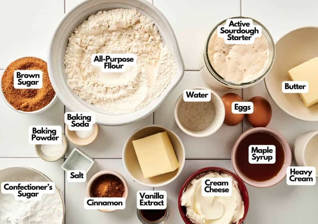Gemini said
An overhead, flat-lay shot of pre-measured ingredients for Air Fryer Sourdough Cinnamon Rolls, including a large bowl of all-purpose flour, a jar of active sourdough starter, brown sugar, two eggs, sticks of butter, and small bowls of maple syrup, cream cheese, heavy cream, confectioner's sugar, cinnamon, vanilla extract, water, baking powder, baking soda, and salt, all arranged on a white tiled background.