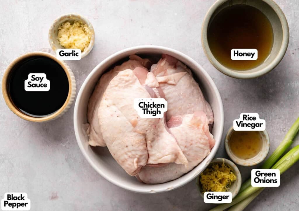 A top-down shot of raw ingredients for Air Fryer Sticky Chicken Thighs arranged on a textured grey surface. The bowl in the center contains four skin-on chicken thighs, surrounded by small bowls of soy sauce, honey, minced garlic, grated ginger, rice vinegar, and black pepper, with two whole green onions on the side.