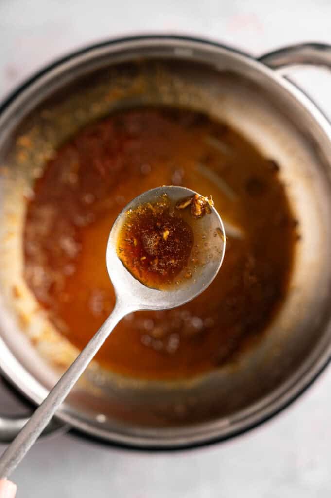 A silver spoon lifts one spoonful of thick, simmered marinade from a stainless steel saucepan, showing visible bits of minced garlic and grated ginger as it thickens over medium heat.