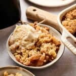 A bowl of Air Fryer Apple Dump Cake with a golden-brown crumble topping and warm cinnamon-spiced apple filling, topped with a scoop of melting vanilla ice cream, served alongside a white ceramic baking dish with three cinnamon sticks on a plate in the background.