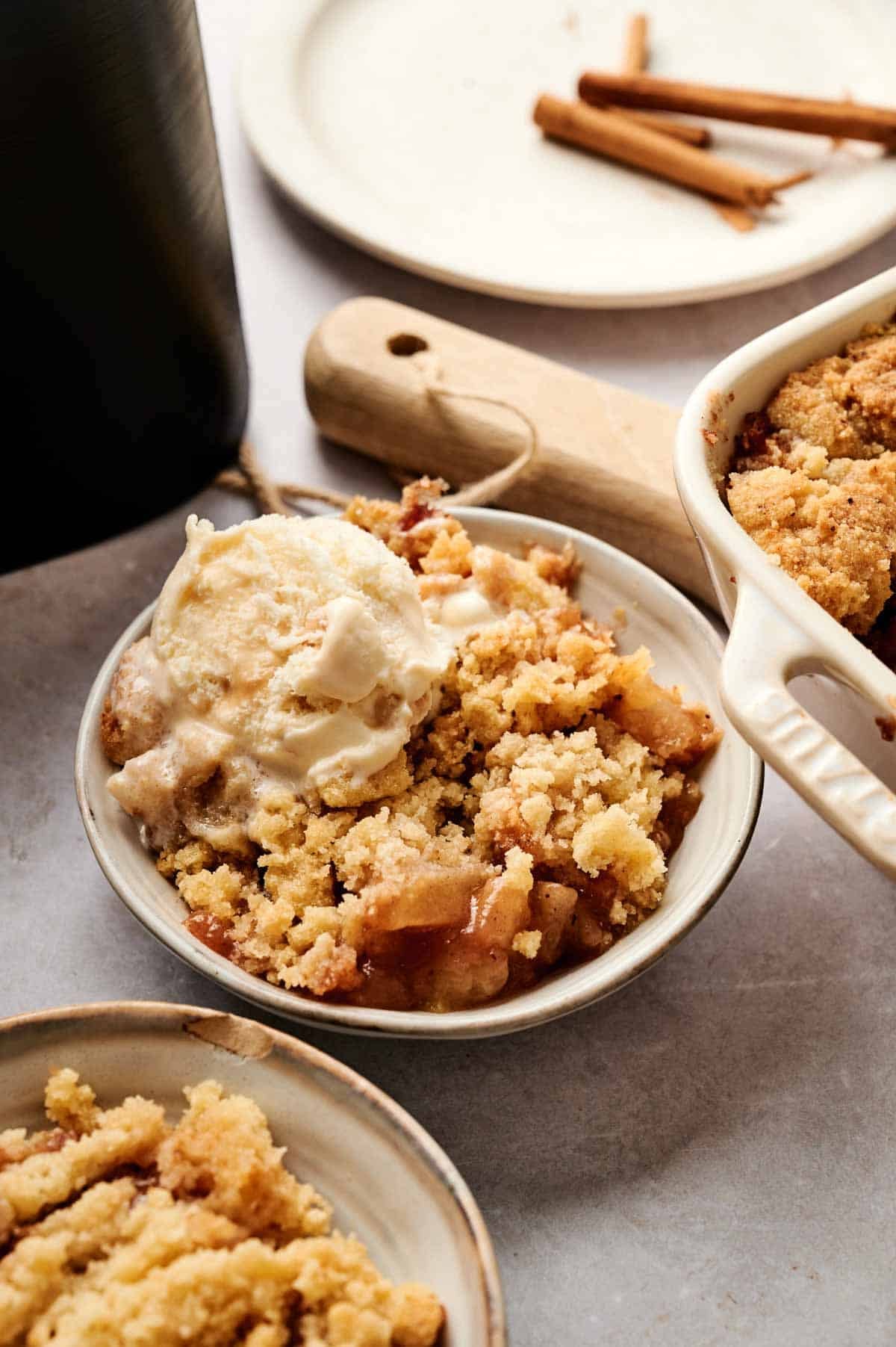 A bowl of Air Fryer Apple Dump Cake with a golden-brown crumble topping and warm cinnamon-spiced apple filling, topped with a scoop of melting vanilla ice cream, served alongside a white ceramic baking dish with three cinnamon sticks on a plate in the background.