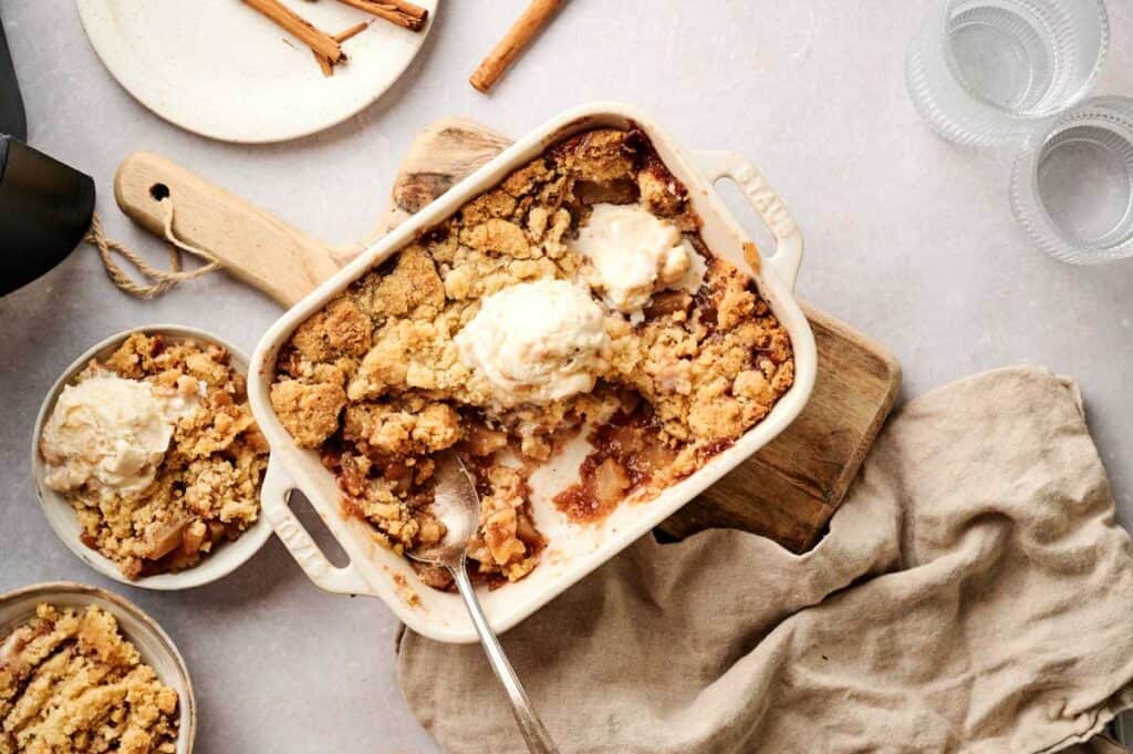 An Air Fryer Apple Dump Cake with a golden crumble topping and melting vanilla ice cream in a white stoneware baking dish, served alongside two smaller bowls and garnished with cinnamon sticks and ribbed glass cups.