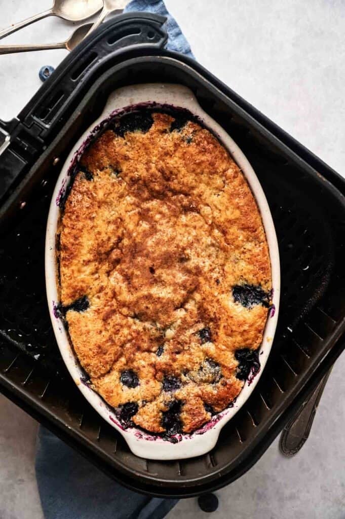 An oval white ceramic dish of Air Fryer Blueberry Cobbler with a golden-brown bubbly crust, sitting directly inside a black air fryer basket. The background shows a light grey surface with a blue napkin, two silver spoons, and a few scattered fresh blueberries.