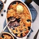 An oval ceramic dish and a small wooden bowl of Air Fryer Blueberry Cobbler with golden-brown crust, each topped with a scoop of vanilla ice cream, served with silver spoons, a blue napkin, a bowl of fresh blueberries, and an air fryer in the background.
