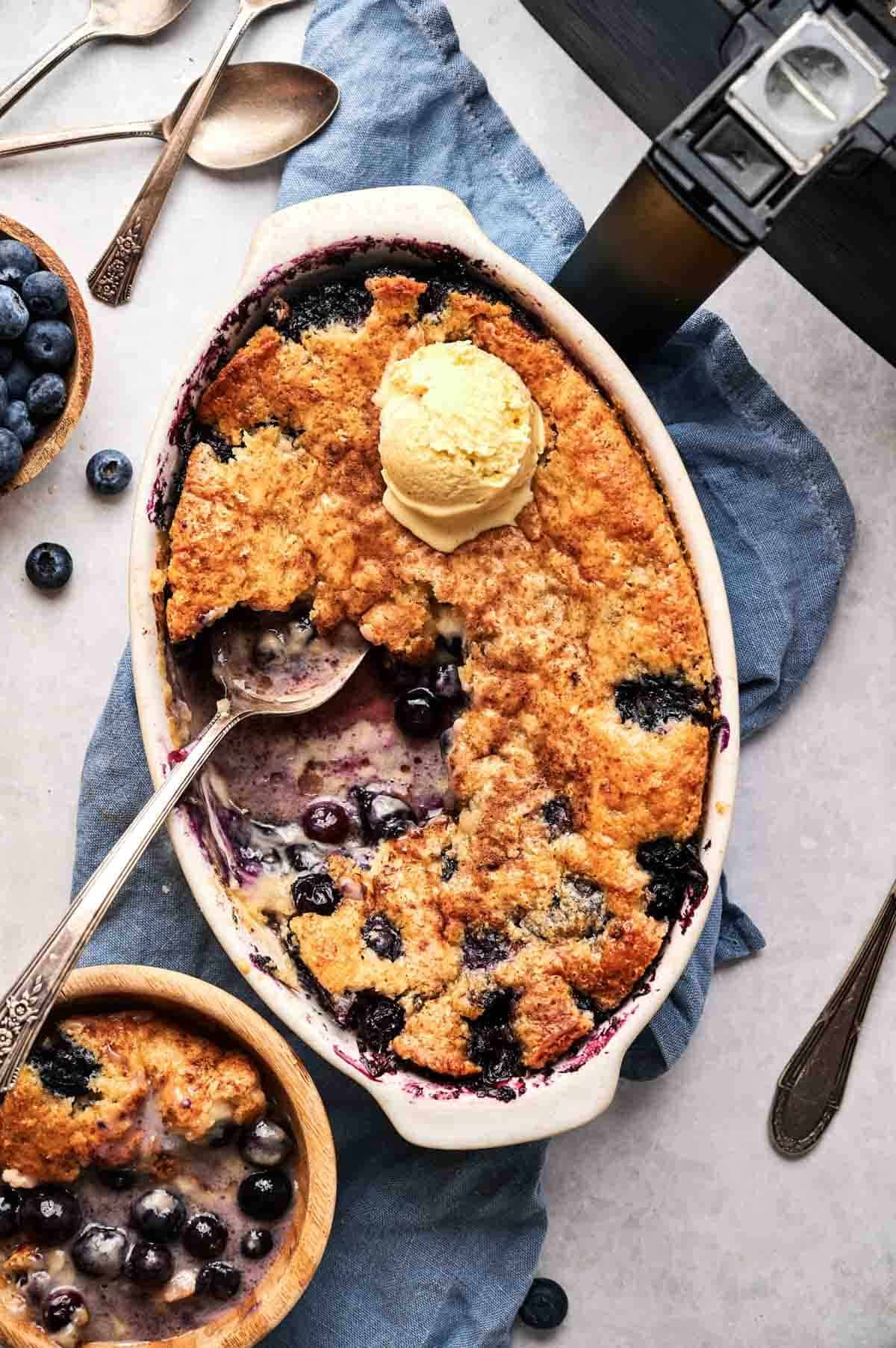 An oval ceramic dish and a small wooden bowl of Air Fryer Blueberry Cobbler with golden-brown crust, each topped with a scoop of vanilla ice cream, served with silver spoons, a blue napkin, a bowl of fresh blueberries, and an air fryer in the background.