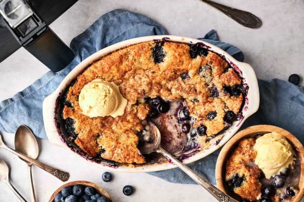 An oval ceramic dish and a small wooden bowl of Air Fryer Blueberry Cobbler with golden-brown crust, each topped with a scoop of vanilla ice cream, served with silver spoons, a blue napkin, a bowl of fresh blueberries, and an air fryer in the background.