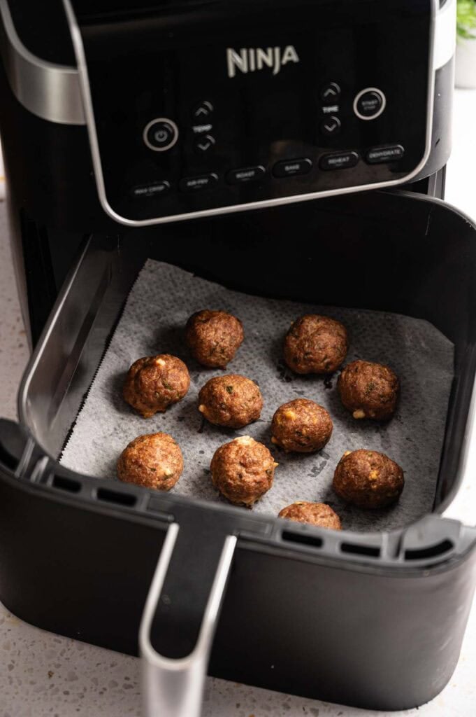 Ten Air Fryer Lamb Meatballs arranged on a sheet of parchment paper inside a black Ninja air fryer basket, showing a browned exterior with visible flecks of herbs and feta cheese.