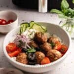 A bowl of seven Air Fryer Lamb Meatballs served over white rice with tzatziki, sliced cucumbers, red onions, cherry tomatoes, and black olives, garnished with fresh dill and mint, alongside small bowls of extra tomatoes, onions, herbs, and a sprig of mint in a glass jar.
