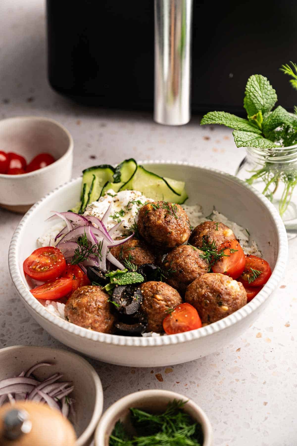 A bowl of seven Air Fryer Lamb Meatballs served over white rice with tzatziki, sliced cucumbers, red onions, cherry tomatoes, and black olives, garnished with fresh dill and mint, alongside small bowls of extra tomatoes, onions, herbs, and a sprig of mint in a glass jar.