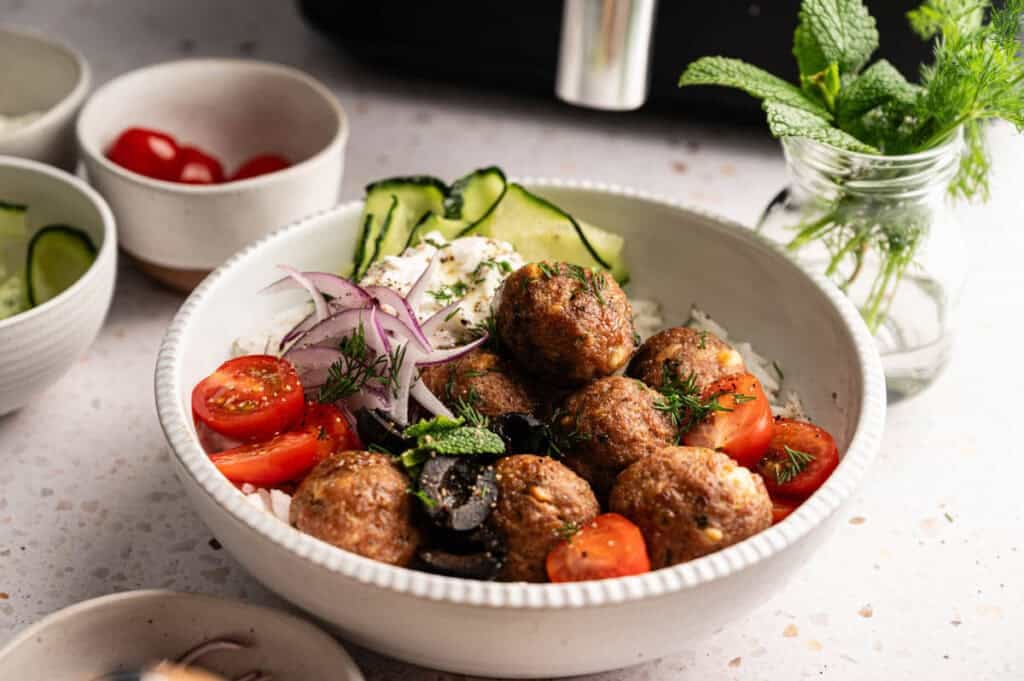 A close-up of an air fryer lamb meatballs being dipped into a small bowl of white tzatziki sauce, with a bowl of eight meatballs, a lemon wedge, and a whole cucumber in the background.
