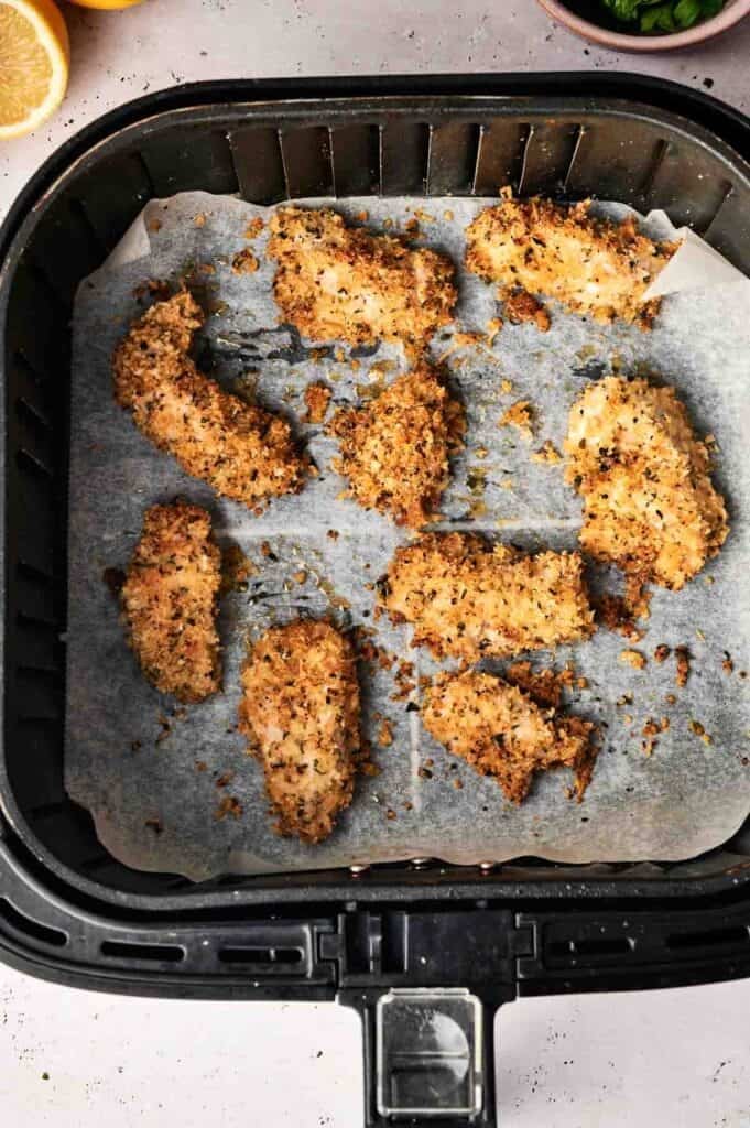 Eight pieces of Air Fryer Parmesan Crusted Chicken arranged on parchment paper inside a black air fryer basket, with a lemon half and a small bowl of fresh herbs visible in the background.