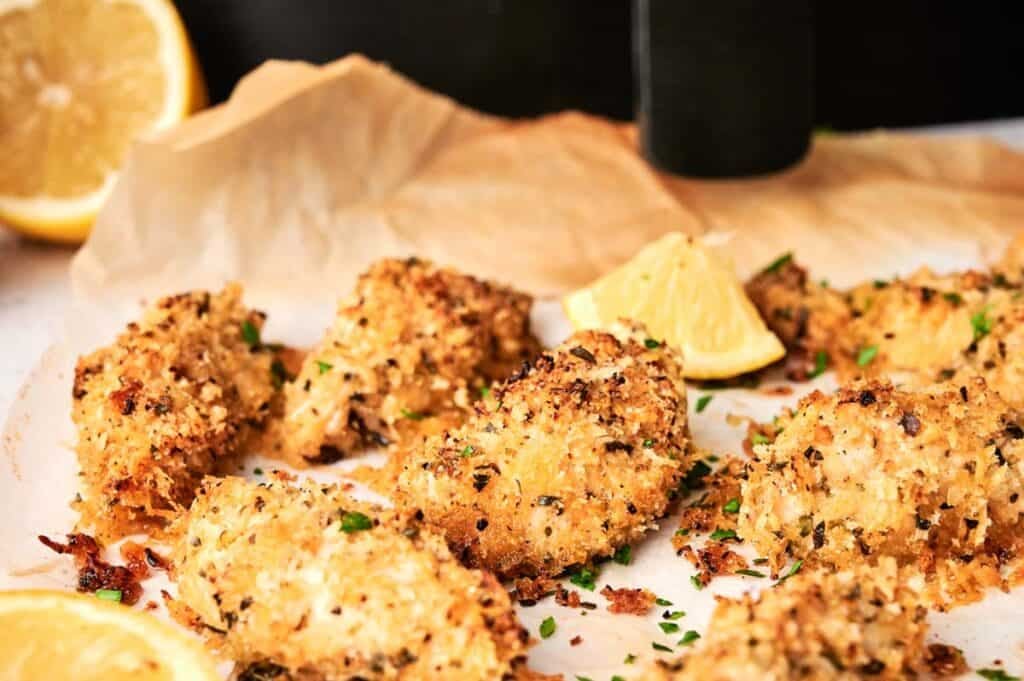 Six pieces of golden-brown Air Fryer Parmesan Crusted Chicken on parchment paper, garnished with fresh chopped parsley and a lemon wedge, with a black pepper grinder in the background.