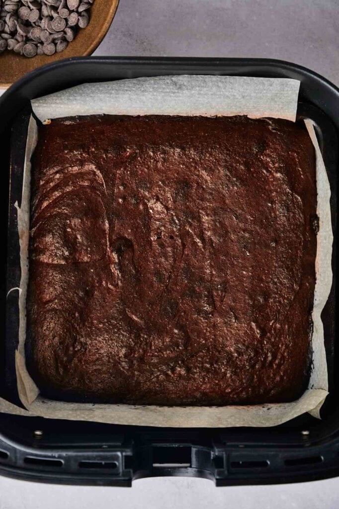 A freshly baked square sheet of Air Fryer Slutty Brownies with a glossy, cracked top, resting in a black air fryer basket lined with white parchment paper. A wooden bowl of chocolate chips is visible on a grey background at the top left.