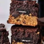 Two stacked Air Fryer Slutty Brownies displaying layers of chocolate chip cookie, a whole Oreo, and fudgy brownie, served on white parchment paper with chocolate crumbs and a blurred black air fryer in the background.