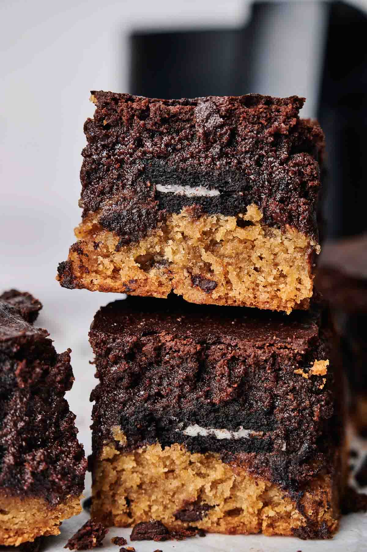 Two stacked Air Fryer Slutty Brownies displaying layers of chocolate chip cookie, a whole Oreo, and fudgy brownie, served on white parchment paper with chocolate crumbs and a blurred black air fryer in the background.