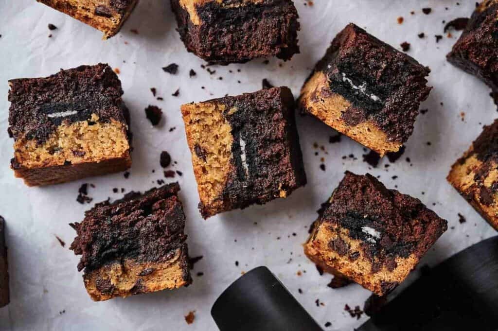 Eight square Air Fryer Slutty Brownies arranged on white parchment paper, showing layers of chocolate chip cookie dough, Oreos, and fudgy brownie, with chocolate crumbs scattered around and a black air fryer basket handle in the foreground.
