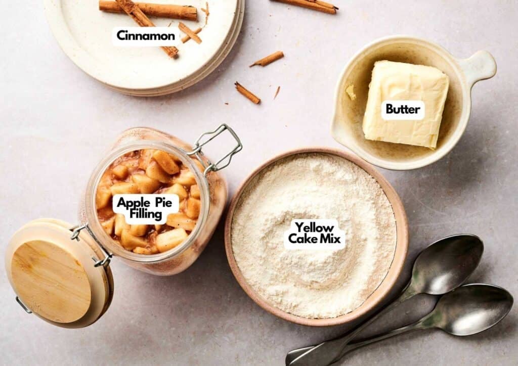 Four essential ingredients for an Air Fryer Apple Dump Cake arranged on a grey surface: a glass jar of apple pie filling, a bowl of yellow cake mix, a dish with a block of butter, and a stack of white plates with cinnamon sticks, all marked with white text labels.