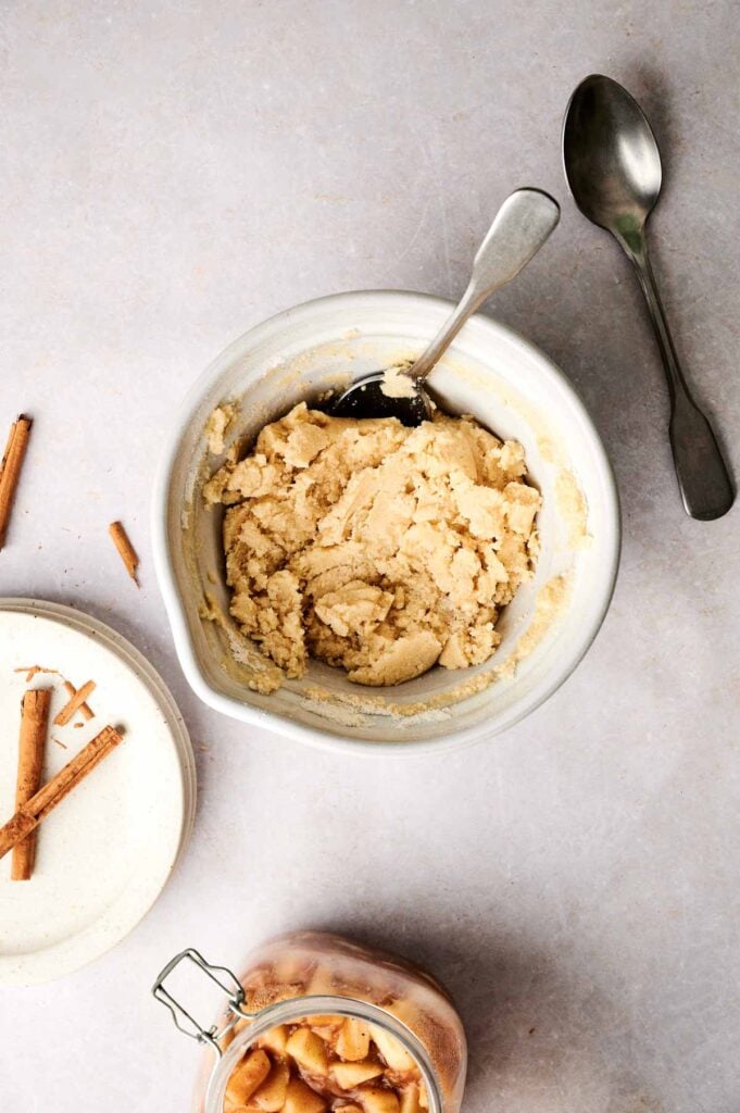 A white mixing bowl containing a thick, pale crumble dough for an Air Fryer Apple Dump Cake with a silver spoon inside, served alongside a glass jar of cinnamon-spiced apple filling, a stack of white plates with two cinnamon sticks, and an extra spoon on the side.