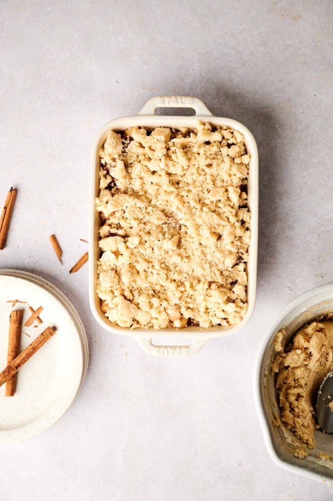 A raw Air Fryer Apple Dump Cake assembled in a white ceramic baking dish with a full layer of pale crumble topping, served next to a plate with two cinnamon sticks and a bowl with a spoon.