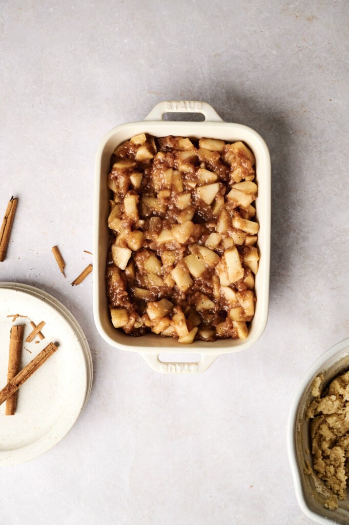 A white ceramic baking dish filled with a single layer of cinnamon-spiced apple filling for an Air Fryer Apple Dump Cake, placed on a light grey surface next to a stack of white plates with two cinnamon sticks and a mixing bowl containing crumble dough.