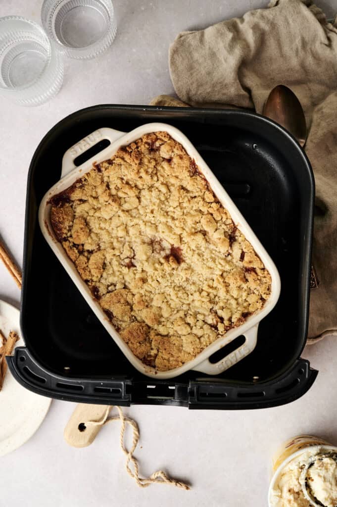 A golden-brown Air Fryer Apple Dump Cake in a white ceramic baking dish, placed inside a black air fryer basket, served alongside two glass ramekins, a plate with a cinnamon stick, a tan linen cloth, and a pint of vanilla ice cream.