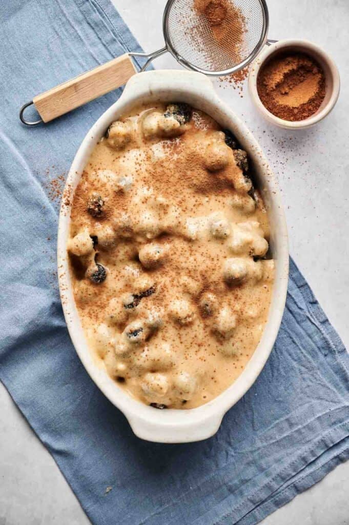 An oval white ceramic dish filled with unbaked Air Fryer Blueberry Cobbler batter, topped with a generous dusting of ground cinnamon. The dish sits on a blue linen napkin next to a small bowl of cinnamon and a metal fine-mesh strainer used for dusting.