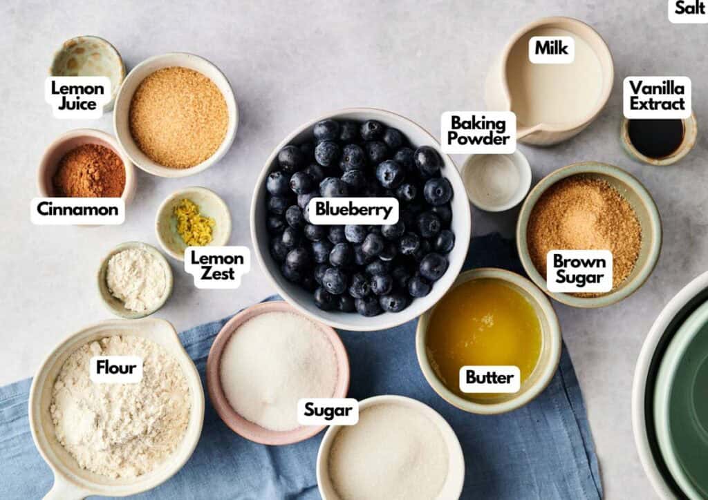 Flat lay of ingredients for Air Fryer Blueberry Cobbler including a large white bowl of fresh blueberries, two bowls of sugar, flour, brown sugar, melted butter, milk, and lemon juice. Small prep bowls contain cinnamon, lemon zest, baking powder, and vanilla extract, arranged on a light grey surface with a blue napkin and stacked mixing bowls in the background.