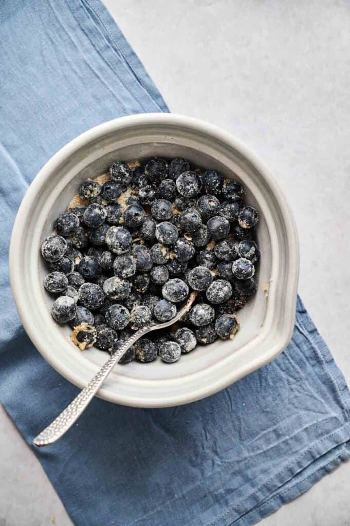 
Gemini said
A light grey ceramic bowl of fresh blueberries for Air Fryer Blueberry Cobbler, tossed in a coating of sugar and lemon zest with a silver spoon resting inside, set on a blue linen napkin against a light stone background.