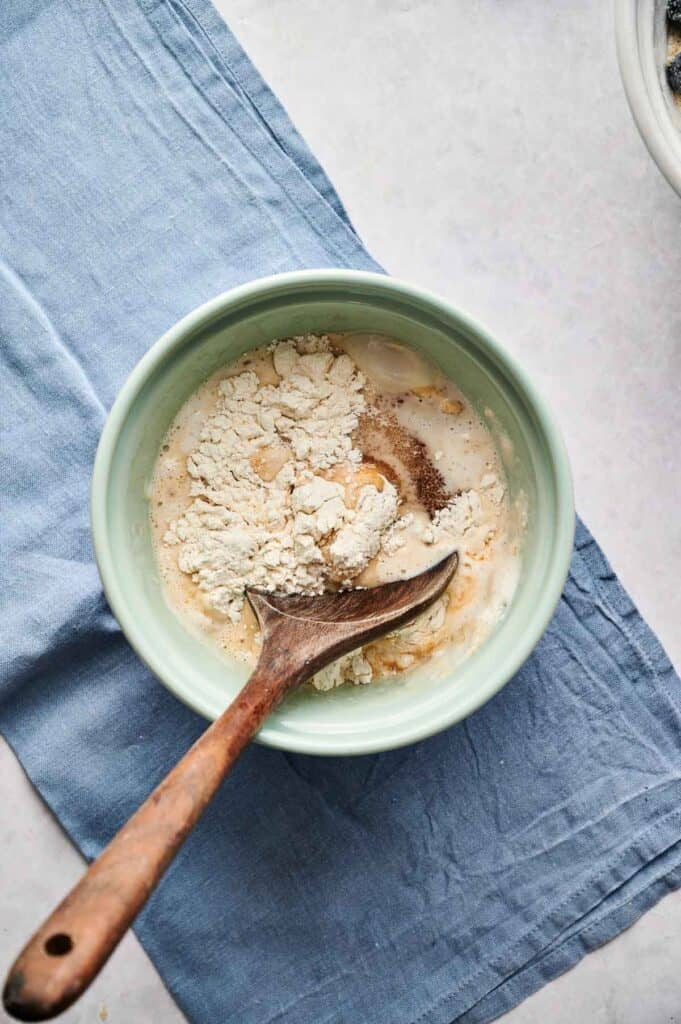 A light green ceramic bowl containing flour and wet ingredients for Air Fryer Blueberry Cobbler batter, being mixed with a wooden spoon. The bowl sits on a blue linen napkin against a light grey surface, with a portion of the blueberry bowl visible in the top right corner.