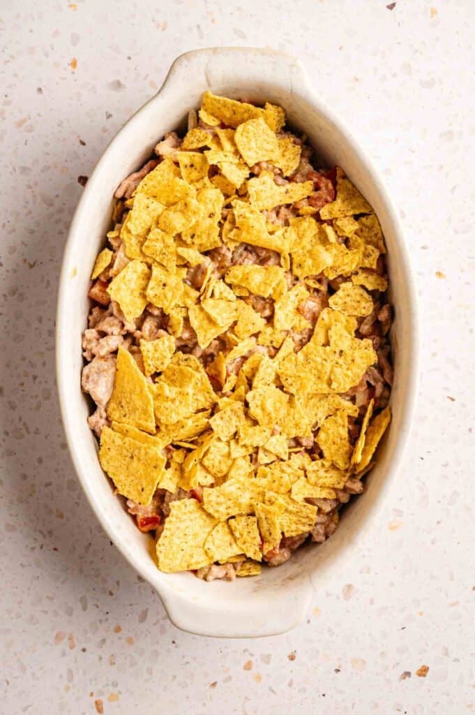 An oval ceramic dish filled with Dorito Casserole, featuring a creamy beef and tomato base topped with a thick layer of crushed yellow nacho cheese Doritos, set against a white speckled countertop.