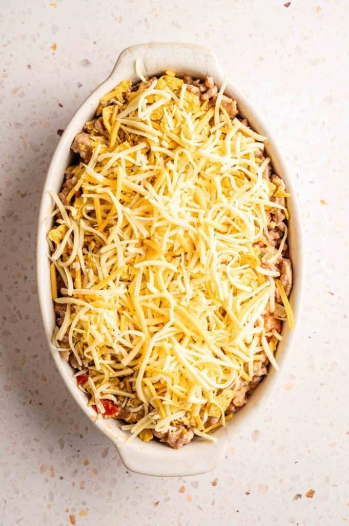 An oval white baking dish filled with Dorito Casserole, featuring layers of seasoned ground meat, crushed nacho cheese chips, and a thick topping of shredded cheddar and Monterey Jack cheese, set against a light speckled stone background.