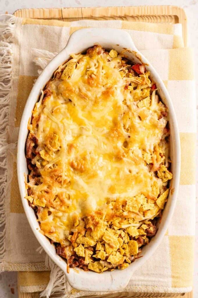 An oval white baking dish containing a Dorito Casserole with a bubbling, melted cheese crust and crushed golden chips, served on a yellow checkered cloth napkin atop a wooden board.