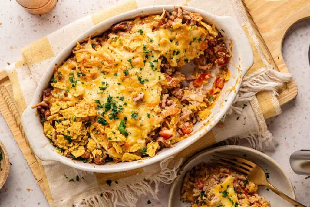An oval baking dish of Dorito Casserole topped with melted cheese, crushed chips, and fresh parsley, served with one plated portion and a gold fork on a yellow checkered linen, with a wooden pepper mill and a small bowl of herbs in the background.