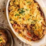An oval baking dish of Dorito Casserole, topped with melted cheese, crushed Doritos, and fresh parsley, resting on a yellow checkered linen, with a small side plate and a gold fork visible in the corner.